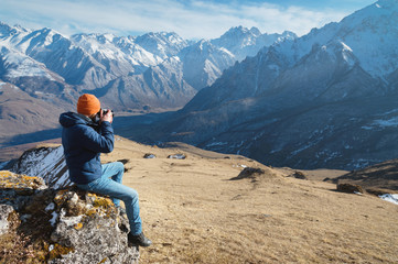 Portrait of a bearded male photographer in sunglasses and a warm jacket with a backpack sits on a big stone and takes pictures on camera against the background of snow-capped mountains