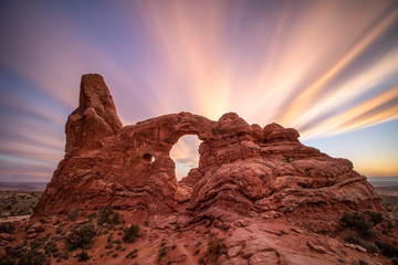 Vibrant pink and orange clouds streaking across the sky behind a naturally formed sandstone arch. Turret Arch, Arches National Park Utah. 