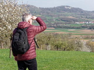 Man in his seventies wearing glasses and carrying a black backpack standing in a meadow, looking into the distance shielding his eyes from the sun with his hands, back view.