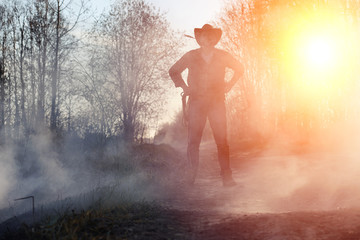 Naklejka na ściany i meble A man is wearing a cowboy hat and a loso in the field. American farmer in a field wearing a jeans hat and with a lasso in the smoke of a fire. A man walks through a burning field