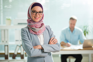 Beautiful young working woman in hijab, suit and eyeglasses standing in office, smiling. Portrait...
