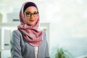 Beautiful young working woman in hijab, suit and eyeglasses standing in office, smiling. Portrait...