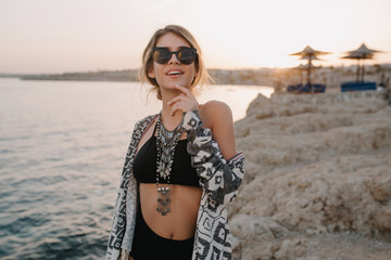 Closeup portrait of smiling young girl on beach, sunset time. Wearing trendy black swimsuit, bikini, stylish sunglasses, necklace, cardigan, cape with ornaments.