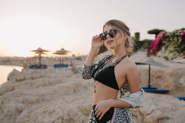 Closeup portrait of gorgeous young girl with slim body wearing stylish black swimsuit, necklace, cardigan, cape with ornaments. Beach, rocks, sunset on background.