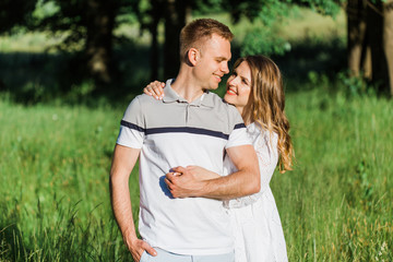 Young pretty couple in love standing in park. Handsome cheerful blonde girl in white dress hugging her boyfriend. Man and woman having fun outdoors