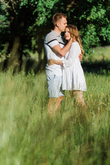 Fototapeta premium Young pretty couple in love standing in park. Handsome cheerful blonde girl in white dress hugging her boyfriend. Man and woman having fun outdoors