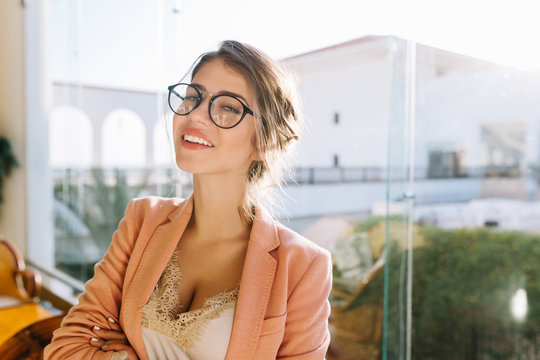 Closeup portrait of young woman wearing stylish glasses, smart lady in elegent pink jacket with beige blouse, cute student. Big window with nice view on background.