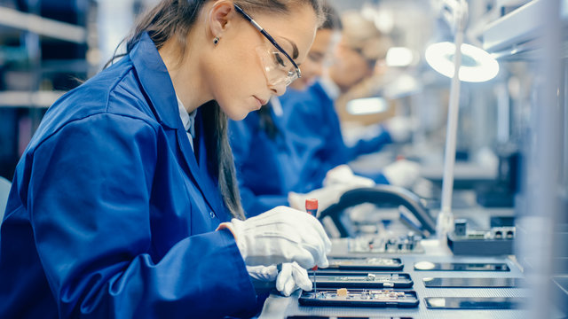 Female Electronics Factory Workers in Blue Work Coat and Protective Glasses Assembling Printed Circuit Boards for Smartphones with Tweezers. High Tech Factory with more Employees in the Background. 