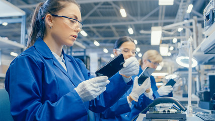 Female Electronics Factory Worker in Blue Work Coat and Protective Glasses Takes a Smartphone...
