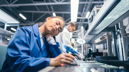 Shot of a Tired Sleeping Female in Blue Work Coat at Her Working Place in Electronics Factory. High Tech Factory Facility with more Employees in the Background.