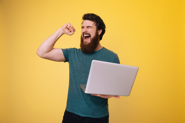 Excited bearded guy holding a laptop while celebrating with rised arm, over yellow background