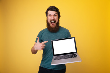 Photo of excited bearded man, screaming and pointing at laptop screen over yellow background
