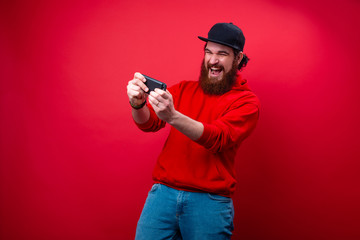 Excited man with beard, playing in mobile phone, over red background
