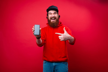 Photo of a bearded guy in hoody and hat, pointing at smartphone and looking at camera over red...