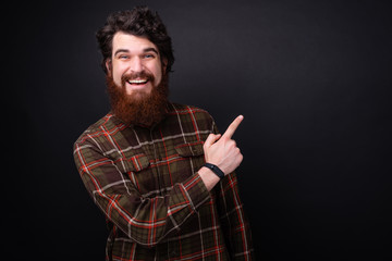 Portrait of bearded man looking at camera while poiting away with finger over darg isolated background