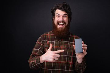 Photo of excited bearded guy pointing at mobile phone and looking at camera over dark background