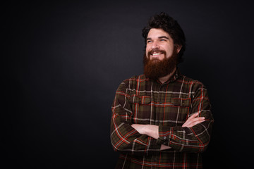 Cheerful bearded man with corssed hands standing over dark wall and looking away