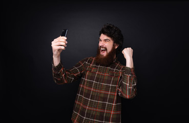 Photo of bearded youn man is celebrating a online prize, screaming  over dark wall