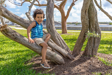 The toddler rests on the tree trunk smiling at the camera. A small boy sits on the base of a curved tree trunk holding on as his dangles.