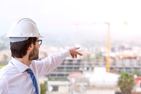 Giovane ingegnere edile con occhiali da vista, caschetto e camicia bianco, cravatta blu. Di fronte a un cantiere edile con gru e palazzina in costruzione visto di spalle.