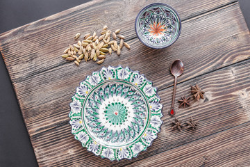Fototapeta na wymiar Authentic middle east style plate, bowl and a bronze spoon with a cardamom and a cinnamon on the wooden desk with a black background. Flat lay, top view. Horizontal orientation.