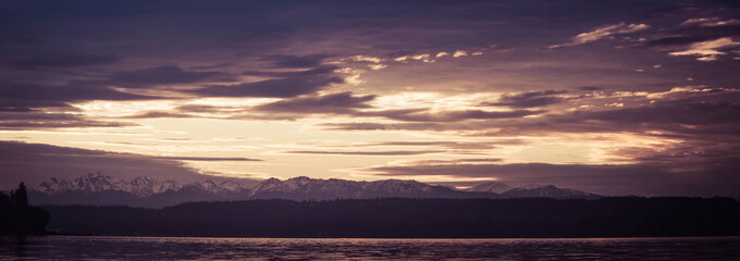A panorama shot on the Pacific northwest with water in the foreground, an evergreen forest in the middle of the image with distant snow covered mountains.  The clouds in the sky are brilliantly lit fr