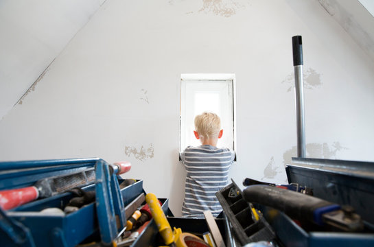 Boy looking out of window in attic to be renovated with toolbox in foreground