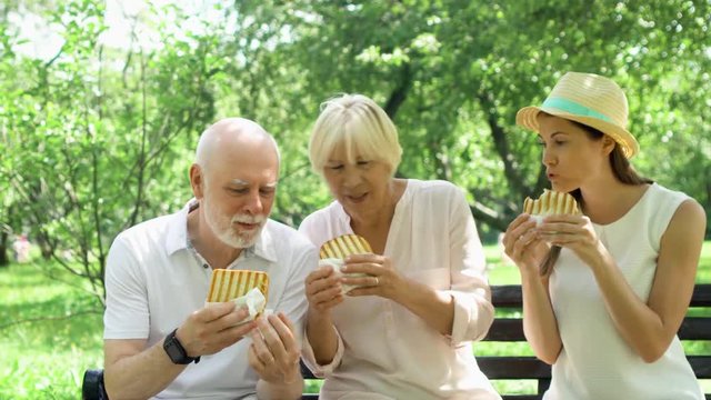 Senior couple with young teenage daughter sitting on bench and eating grilled sandwich with ham in public park. Hungry tourists family having lunch in garden enjoying summer sunny day