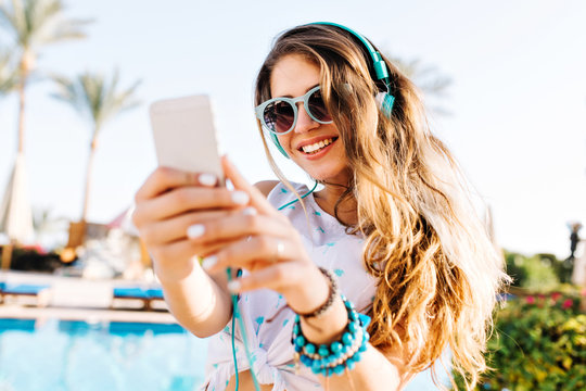 Smiling long-haired girl in sunglasses and earphones making photo of beautiful landscape with exotic palm trees. Joyful tanned young woman with phone chilling out near the pool in sunny morning