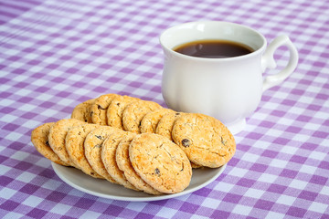 White porcelain cup with tea and sweet oatmeal cookies with pieces of dark chocolate on a white saucer on a white purple checkered tablecloth.