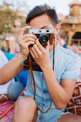 Portrait of boy with short black hair, holding retro camera and making photo, while sitting in open-air cafe. Young man in denim light-blue shirt with trendy hairstyle taking picture of photographer