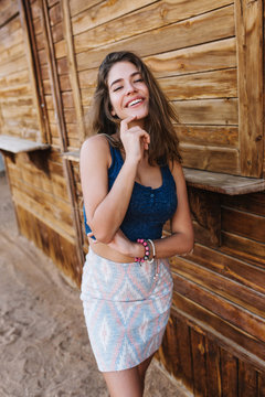 Happy slim girl in trendy skirt and blue tank-top laughing and touching her chin. Portrait of adorable tanned young woman with beautiful hairstyle having fun outside near the wooden stall.