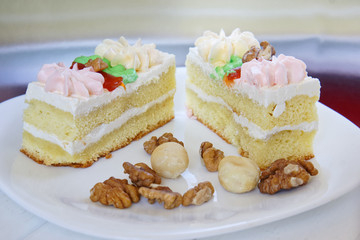 Sponge cakes with cream flowers, soaked in brandy, lie in a plate next to scattered macadamia nuts and walnuts on a light background. Wooden table. Close-up