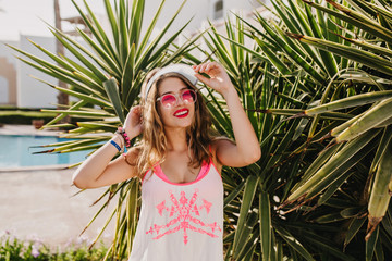 Portrait of laughing long-haired girl with amazing suntan wearing white hat and trendy tank-top. Joyful young woman in sunglasses and stylish accessories posing with smile standing near palm trees