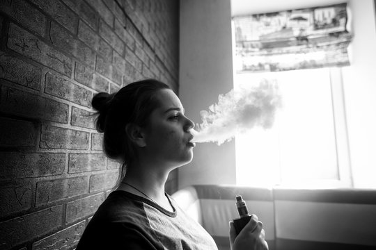 Vape teenager with  problem skin. Portrait of young cute girl smoking an electronic cigarette in the bar. Bad habit that is harmful to health. Vaping activity. Black nad white.