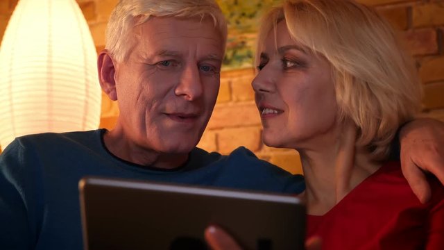 Closeup shoot of aged happy couple using the tablet sitting on the sofa resting together indoors in a cozy flat