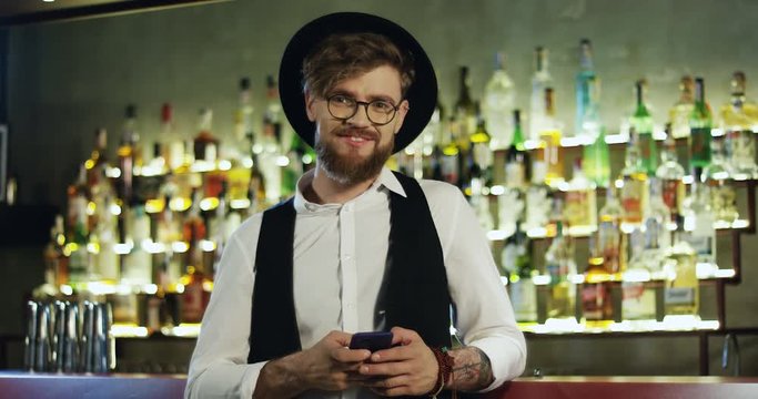 Young stylish and handsome Caucasian man in hat and glasses tapping, scrolling and texting on the tablet computer, then smling cheerfully to the camera at the bar.