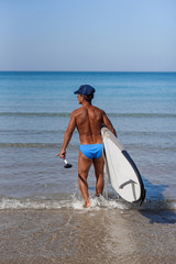 Naklejka na ściany i meble Tanned man on the beach of the sea with a surfboard and oar in his hands goes into the water. The concept is a sporty and healthy lifestyle.