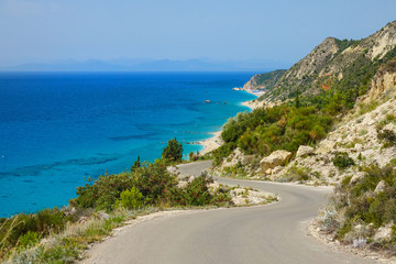 Empty asphalt road winds along rugged Mediterranean terrain and turquoise ocean.