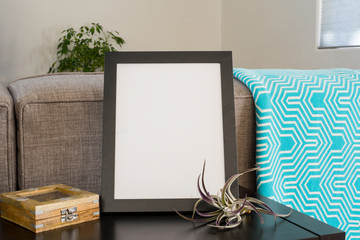Black 8x10 frame on a livingroom  table top with a teal blanket airplant and puzzle box.