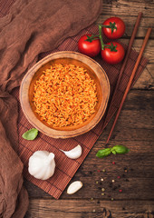 Wooden plate bowl of rice with tomato and basil and garlic and chopsticks on brown bamboo place mat on wooden background with dark cloth. Top view.