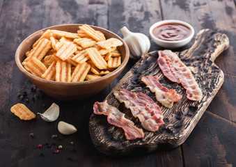 Bacon flavoured snacks chips with grilled bacon rashers on vintage chopping board with garlic and sauce on wooden background.