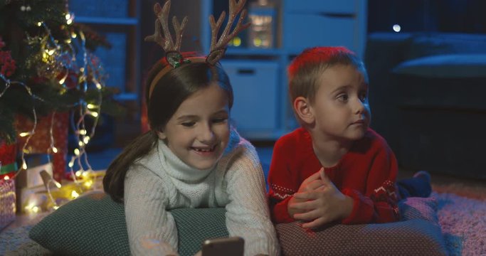 Close up of the small happy sister and brother lying on the floor at the Christmas tree and watching cartoons on the smartphone in the dark room.