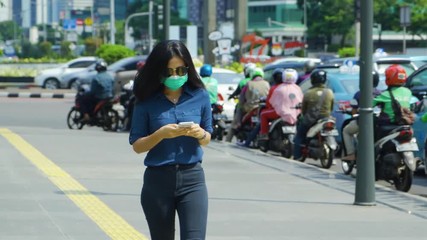 JAKARTA, Indonesia - April 24, 2019: Air Pollution Concept. Young woman wearing a mask while walking on the sidewalk and using a mobile phone. Shot in 4k resolution