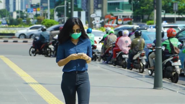 JAKARTA, Indonesia - April 24, 2019: Air Pollution Concept. Young woman wearing a mask while walking on the sidewalk and using a mobile phone. Shot in 4k resolution