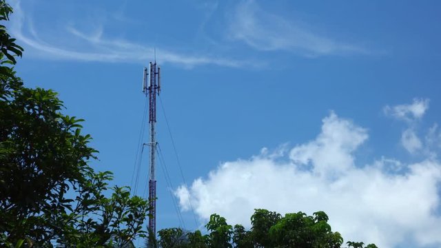 Mobile communication mast in red and white time lapse with treetops and deep blue tropical cloudy skies. Network broadcasting tower.
