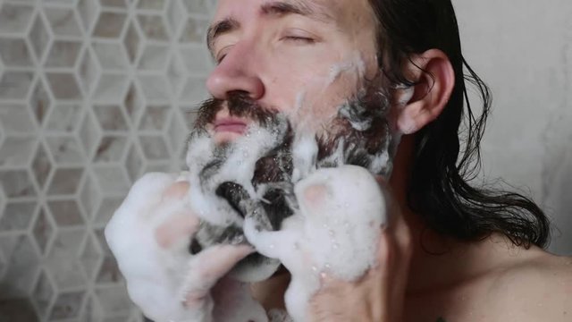 Portrait of young man is washing his beard with shampoo and foam in the shower enjoying the process with closed eyes, close-up.