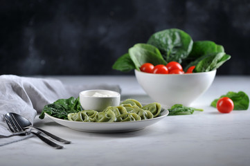 Ravioli with spinach and sour cream. In the background is a bowl of spinach and cherry tomatoes. Close-up. Dark background.
