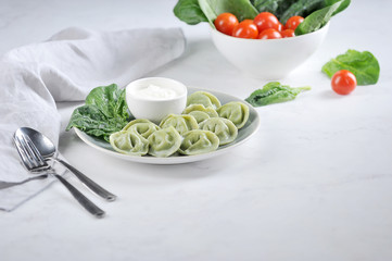 Ravioli with spinach and sour cream. In the background is a bowl of spinach and cherry tomatoes. Light background. Free space to place text.