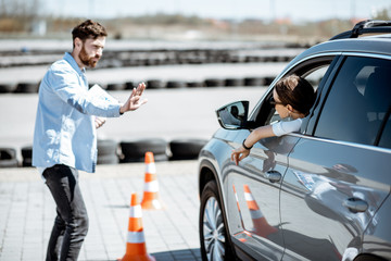 Male instructor teaching young woman driver to park a car on the training ground with traffic cones at the school